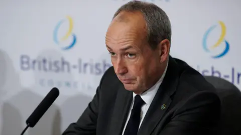 Lyndon Farnham is dressed in a dark suit, with a black tie. He has a microphone in front of him and is looking serious.