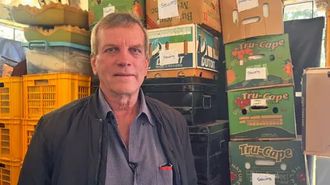 Deon Theron in a shirt and jacket stands in front of boxes and crates.