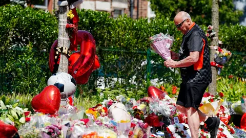 PA Media A man in a black t-shirt, black shorts and sunglasses stands amid a sea of tributes including flowers, football shirts, balloons and flowers with a bouquet in his hand
