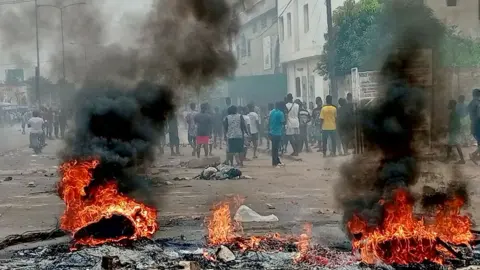 As barricadas improvisadas da Reuters queimam como as pessoas protestam (suas costas para a câmera) contra o líder de longa data do Togo, Faure GNASSINGBÉ, em Lomé, Togo - 26 de junho de 2025