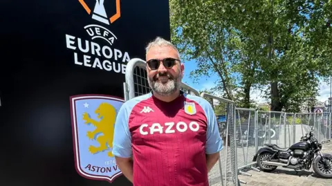Jamieson Hill stands with his hands behind his back. He is wearing an Aston Villa home shirt, which is predominately claret with light blue sleeves. 