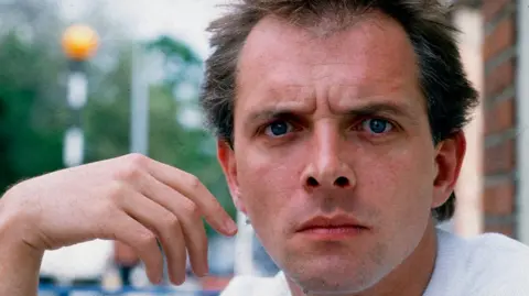Rik Mayall, a clean-shaven man with dark hair, looks at the camera, glaring at the camera in a light-hearted way. His hand is raised and hangs limp by the side of his face.