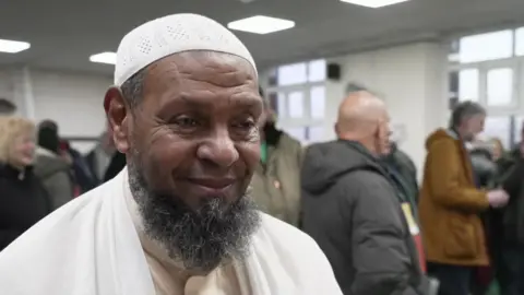 An imam with a long beard and a white prayer cap stands smiling in a room full of people.