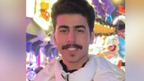 Cambridgeshire Police/Family Mohammed Algasim: a dark-haired man with a dark moustache. He is wearing a white anorak-style coat and standing in front of a multi-coloured background 
