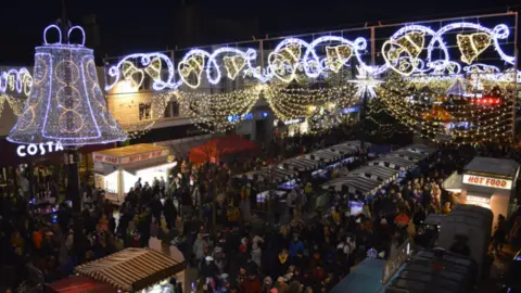 Charnwood Borough Council A packed town centre at night with bright Christmas lights, including a huge bell of lights above the crowds