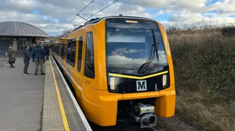 One of the new Metro trains which is bright yellow with black accents. People are waiting on the platform beside the train.
