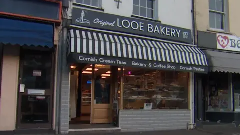 Looe Bakery with a sign saying its name in white on a black background. It has a black and white pattern of stripes above it and also says Cornish Cream Teas, Bakery & Coffee Shop and Cornish Pasties.