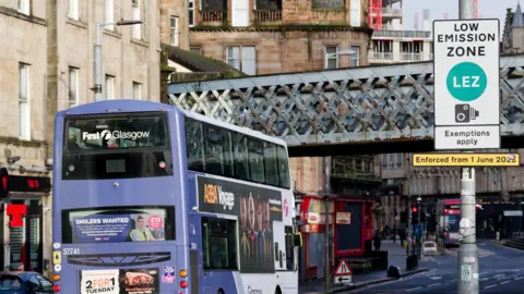 A bus approaches a railway bridge with a low emission zone sign on a lamp post in the foreground
