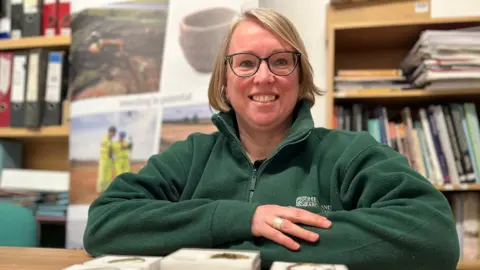 Emma Baugh/BBC Sara Machin sitting with her arms resting flat on a table which has some finds on it