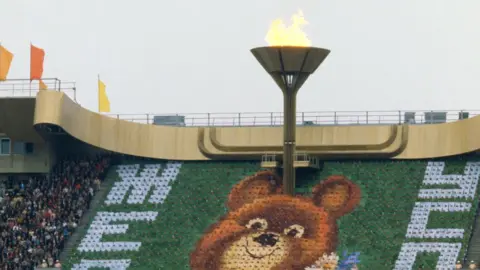 The huge Olympic torch seen at a stadium during the opening ceremony of the Olympic Games in Moscow in 1980.  The seating area shows a big image of the Olympic mascot - a teddy bear known as Misha.
