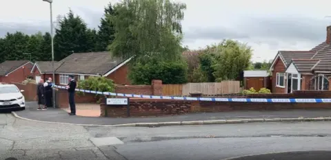 Police tape cordons off an area including Finchley Drive around Chadwick Road with two police officers in attendance as a resident in the street looks on.