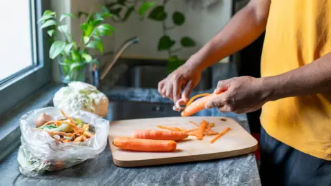 A person peels carrots over a wooden chopping board. They have a bag next to them with the peelings in and with other food waste. In the background there is a sink and plants.