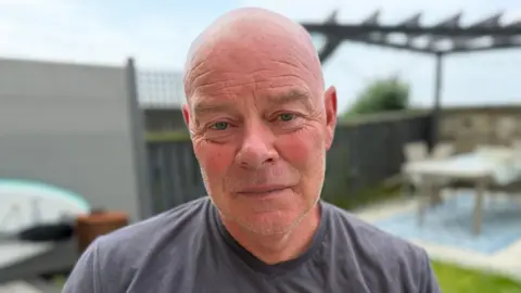 A bald man looks at the camera. He is sitting in a back garden and wears a grey T-shirt