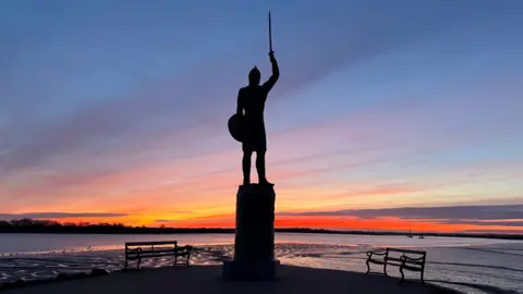 MALDON CHRIS/WEATHERWATCHERS A sunrise behind a silhouette of a statue of 10th-century Anglo-Saxon Ealdorman, Byrhtnoth, who is holding aloft his sword and a shield.
