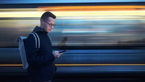 A man standing at a railway station looking at his phone as a blurry image of a train passes him.