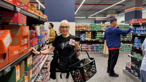 Francisco Velasquez/BBC A woman with glasses in a black shirt and white pants holds a shopping basket