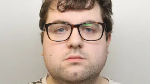 A custody image of Nathan Bennett. He has brown glasses on, a neutral expression, and mid-length brown hair. He is wearing a cream T-shirt with a brown, leaf-life design on it.