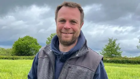 Olly, wearing a blue hoodie and grey gilet, is standing in the middle of a field on a cloudy day. 