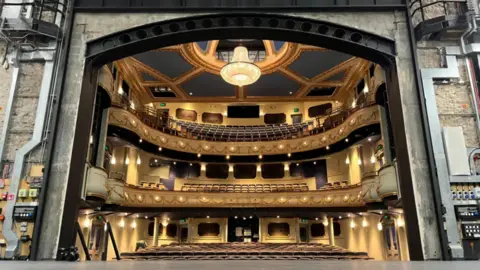 A wide shot of Jersey Opera House. It is a beautiful golden theatre with three tiers of seats. The ceiling is a dark grey interspersed with golden decoration.