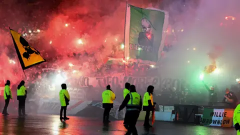 Smoke and red flares visible in a stand full of football fans. Multiple stewards and police officers wearing high vis jackets are visible. Two large flags emerge from the crowd, one is yellow and black with a skull on it and the other is green and with a skeleton on it.