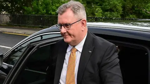 A man with grey hair, glasses gets out of a car. He is wearing a dark grey suit, a white shirt and and orange and white tie.A man with grey hair, glasses gets out of a car. He is wearing a dark grey suit, a white shirt and and orange and white tie.