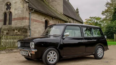 Their Last Mini Adventure A black converted Mini Clubman hearse, viewed from the front at an angle. There is a church behind the vehicle and a graveyard.