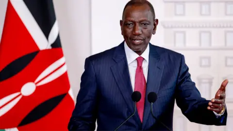 President Ruto in a navy pinstripe suit and pink tie speaking in front of two microphones - with a hand raised in explanation. A Kenyan flag can be seen behind him.