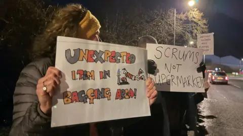 Two people holding signs protesting the Aldi. One says Scrums not supermarkets and the other says Unexpected item in bagging area. 
