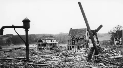 A black and white photograph showing old-fashioned rural German buildings, with timber frames, destroyed in the Dambusters raid, there is debris piled up around the remains of the buildings. 
