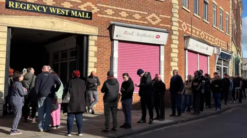 BBC/Eve Connor Dozens of people standing outside on the street, waiting in the queue to get into the record shop. There is a sign above a doorway into the shopping mall that says Regency West Mall. People can be seen entering this entrance, as the queue continues along to the shop. Next door is a shop with two sets of doors or windows protected by candy-pink roller shutters.