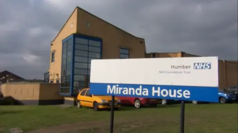 The outside of Miranda House, Hull. The yellow brick building has blue window frames and stands behind a blue and white NHS sign.