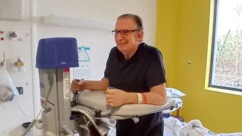 Kevin Pardy A man with black, grey hair stands with the help of a medical support in a hospital room which has a white wall and a yellow wall behind him. He wears half-rimmed glasses and a blue top.