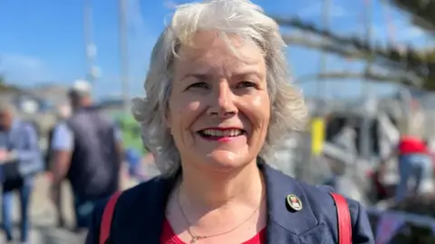 A close up of Sue Aldwell. She has short grey hair and is smiling into the camera. She is wearing a navy jacket and pink top.