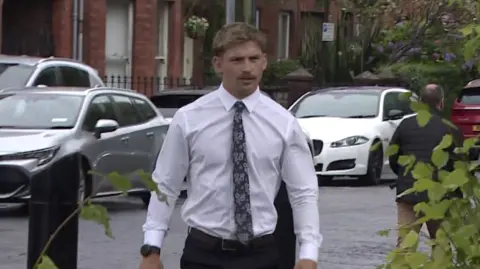 Kavan Keenan is a young man with short blonde/ brown hair and a moustache. He is walking towards the gates into the court as the picture is taken. He is wearing a white long sleeve shirt and a black patterned tie. Behind him is a silver car and two white cars parked along the street in front of red brick terrace houses.