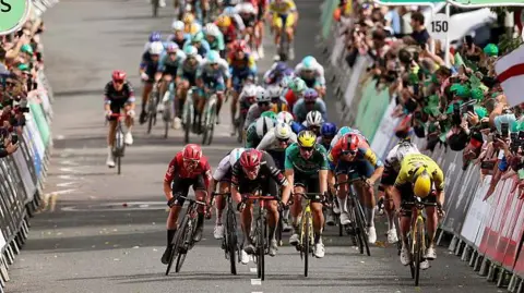 A group of cyclists racing against each other on a road. Members of the public are gathered behind barriers watching. 