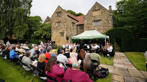 Dozens of people sit on chairs in the garden of the tall stone building. There are watching a brass band playing under a green National Trust gazebo. There are tall, lush green trees in the background.