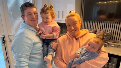 BBC/Jim Scott Liam Jackson, wearing a blue hoodie and holding their two-year-old daughter, stands next to Megan, who is wearing a pink hoodie and is holding their two- month old son, Albie. They are standing in front of pre-packaged cakes.