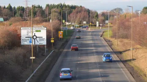 Jim Barton Cars travelling along a road towards a roundabout on the A7. It is a bright day with long shadows and autumnal trees at the side of the road