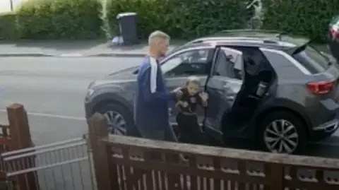 PA Wire A man grabs a child by his arm as they exit a black car. In the foreground you can see a gate attached to a wooden fence leading to a house.