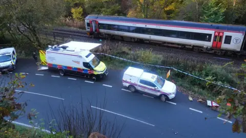 Carro de polícia, van Network Rail e jipe ​​de emergência Network Rail na beira da estrada próximo aos trilhos enquanto o trem acidentado permanece nos trilhos à luz do dia. As portas finais do carro estão abertas e há fita adesiva na frente dos trilhos.