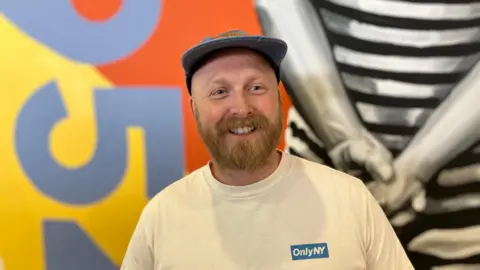A man in a white t-shirt with a blue hat and a ginger beard. he is smiling looking at the camera. behind him is part of a mural he has painted featuring bright colours like orange, yellow and blue.