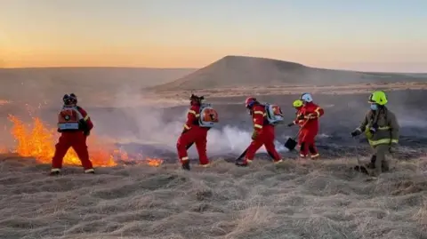 West Yorkshire Fire and Rescue Service Six firefighters in red overalls tackle a fire on a grassy area of moorland 