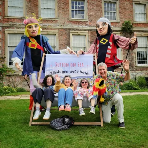 South & East Lincolnshire Councils Partnership Two giant puppets stand at either side of a giant deckchair which has the wording Sutton on Sea wish you were here. There are three women sitting in the giant deckchair, with a man kneeling on the grass to the right of it in the picture. They are all smiling at the camera and three of them are waving.