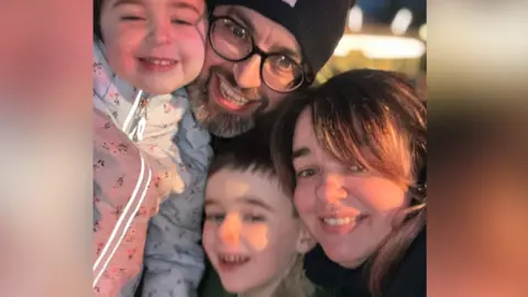 Cillian Kirwan and his wife Louise and two children. They are all grinning at the camera. Cillian is wearing a black beanie, black glasses and his daughter is wearing a white floral coat, his son is grinning next to his mum who has long brown hair. 