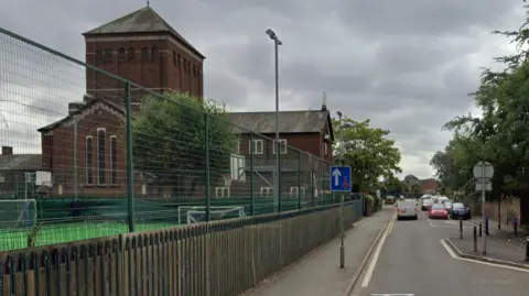 A narrow through road with give way sign, with a large church building/converted primary school to the left 