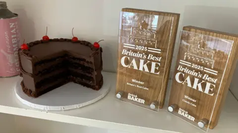 BBC A four layer chocolate cake sits on white shelf next to two wooden awards with glass fronts that read "2025 Britain's best cake"
