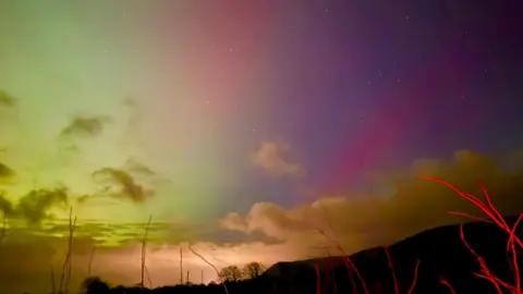 Reed Timmer A sky that turn from green on the left into pink and purple on the right. There is cloud towards the bottom where there is a sillhouette of a headland.