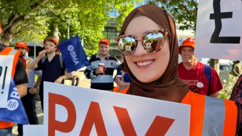 Amanda White/BBC A woman wearing a brown headpiece with cheetah-print sunglasses, smiling and holding up a sign. She is wearing an orange hi-vis top. Behind her are several other people also holding up signs.