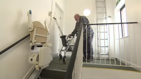 The image shows a stairwell. Chris Duffin is standing on the landing with a walker. He has crutches in the basket of the walker. He is facing a fire door.