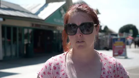 Shaun Whitmore/BBC Eliza Henshall, a woman standing outside in Great Yarmouth. She is looking directly at the camera and has a pair of large dark sunglasses over her eyes. She is wearing a pink floral T-shirt.
